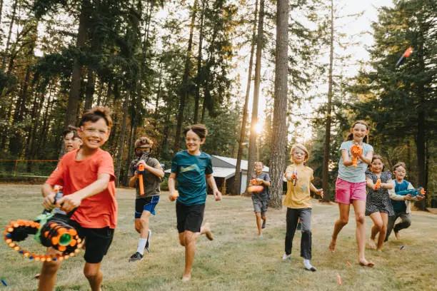 A multiracial group of elementary age and tween boys and girls run and shoot nerf guns toward the camera while having a fun nerf gun battle in a grassy field surrounded by fir trees. The friends are attending a back yard birthday party.