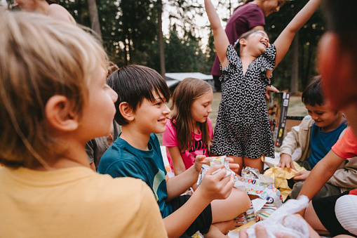 A multiracial tween boy who is having a birthday party outside in the back yard of his home smiles while opening birthday gifts surrounded by his friends.