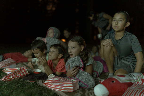 A multiracial group of elementary age and tween kids sit in the grass outside on a warm night and eat popcorn and candy while watching a movie on a projector screen.