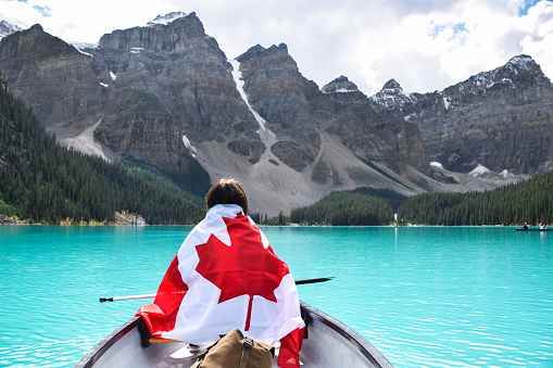 Young girl from behind wrapped in a canadian flag in a canoe in the middle of turqouise blue Moraine lake surrounded by mountains