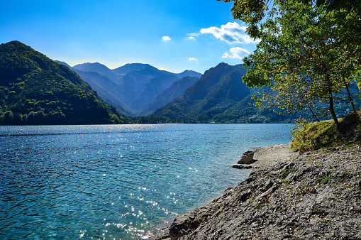 crystal blue water of lake Ledro, stone beach, rocks, mountains, branch, forest, blue sky