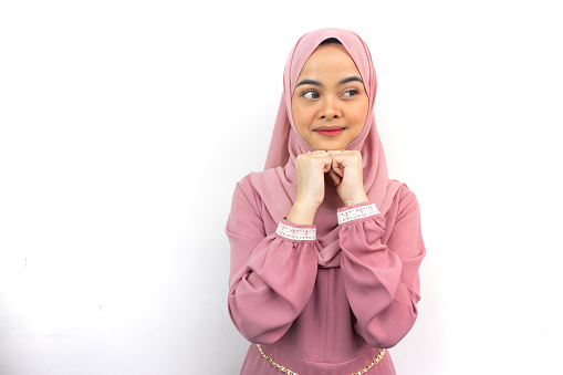 Beautiful smiling Asian woman in pink hijab keeps hands under chin while glanced, remembers pleasant moment isolated on background