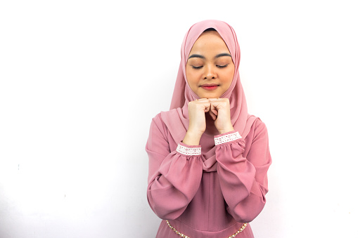 Beautiful smiling Asian woman in pink hijab keeps hands under chin while closing eyes, isolated on background