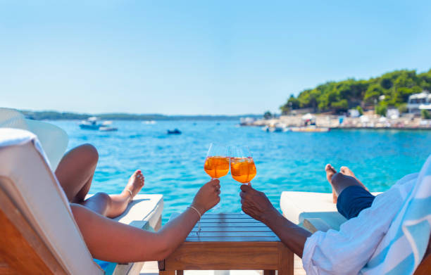 Couple relaxing and toasting with a Spritz cocktail on a beach deck over the ocean. Couple relaxing and toasting with a Spritz cocktail on a beach deck over the ocean. Turquoise ocean is crystal clear.  horizon in the distance. exclusive travel stock pictures, royalty-free photos & images