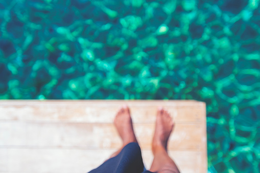 Defocussed image of a Man standing on a jetty by the ocean. Turquoise water is crystal clear. only the mans feet can be seen.