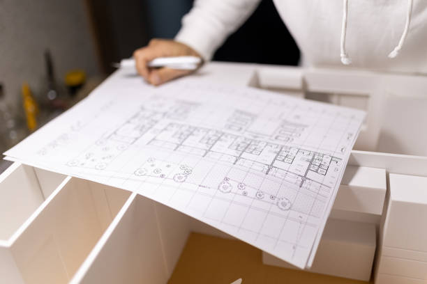 A mid tight clip of a Japanese architect planning the layout of a new house with his blueprints on paper and a large model house.