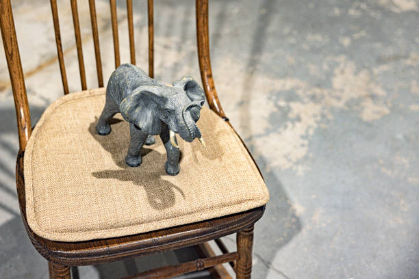 A large toy elephant standing on a cushion on a rocking chair in an industrial floored, but otherwise empty and unfinished basement room. Conceptually portraying the often unspoken \