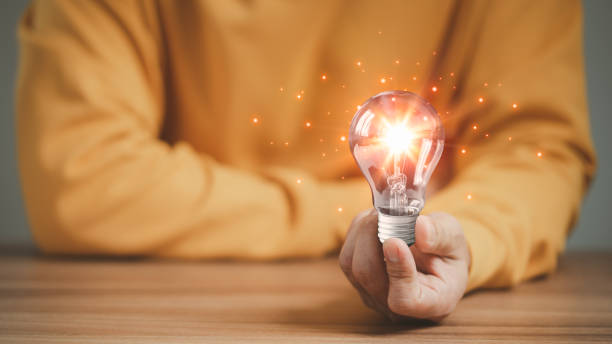 istock Man holding a glowing abstract light bulb on a wooden table, business problem solving ideas and creative marketing ideas Inventing and learning new things. 1454313501