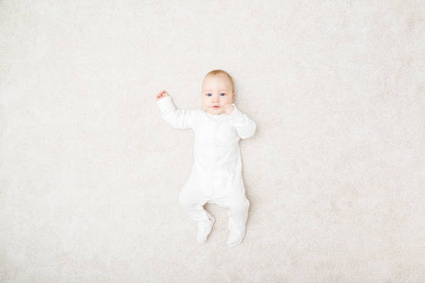 entzückender kleiner junge in weißem body, der auf dem rücken auf hellbeigem heimteppich liegt. draufsicht nach unten. posieren und in die kamera schauen. 5 bis 6 monate altes kleinkind. - carpet floor lying down people stock-fotos und bilder