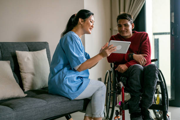 An Asian female healthcare worker teaching Mature Asian disabled Malay man in wheelchair how to online by using digital table at living room. stock photo