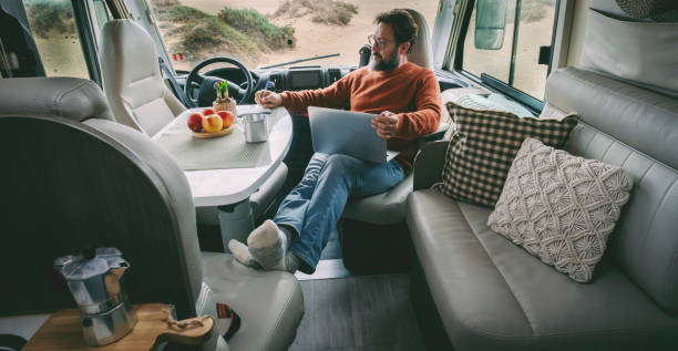 un homme seul assis et se relaxant à l’intérieur d’un camping-car mode de vie alternatif à la maison vanlife hors réseau en utilisant un ordinateur portable et en prenant un café en toute liberté. activité de loisirs de voyage en camping-car int - motor home park camping luxury photos et images de collection