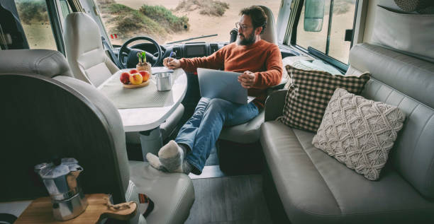 One alone man sitting and relaxing inside a camper van alternative home vanlife lifestyle off grid using a laptop and taking a coffee in total relax freedom. Indoor motor home travel leisure activity stock photo