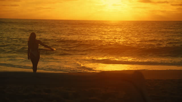 Young surfer woman with long hair is walking from the beach to the sea during sunset,carrying a surfboard underneath her arm