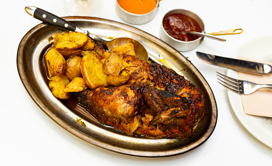 Just cooked roasted chicken served on metal dish with fried potatoes, two sauce and serving pieces.