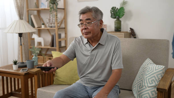 istock Old elderly Asian man happy watching TV self isolating during coronavirus outbreak lockdown in country. relax mature male sitting at home on sofa enjoy comedy television show with joy 1454330475