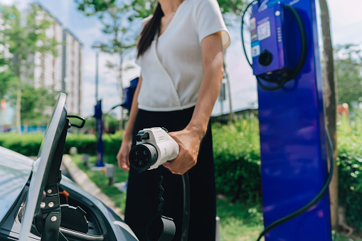 Unrecognizable woman holding an electric charging plug and plugging it into car for refilling fuel.