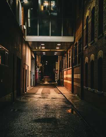 A vertical shot of a dark alley at night with a footbridge above it between old-style brick buildings