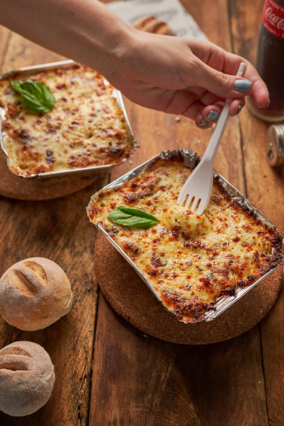 istock Two foil dishes with a baked meal with cheese on top, and a hand holding a plastic fork 1454343717