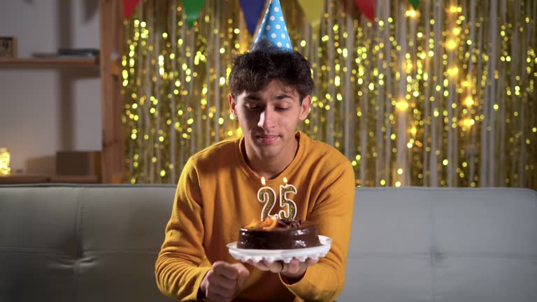 Cheerful arabic man light, blows candles 25 on birthday cake wears cone hat, makes wish, has happy festive mood at decorated home. Festivity