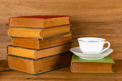Closeup view stack of colorful old books with white ceramic coffee cup on wooden table.