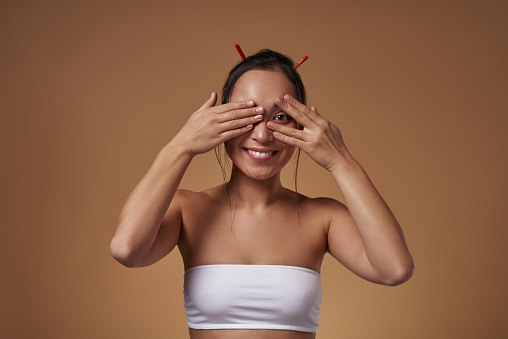 Smiling asian girl covering her ear and looking through fingers at camera. Pretty young brunette slim woman wearing tank top. Female beauty. Isolated on orange background. Studio shoot. Copy space