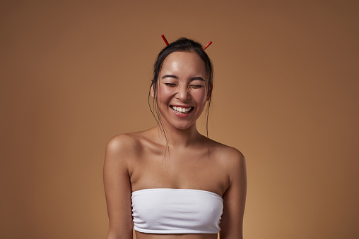 Laughing fashionable asian girl with closed eyes. Excited pretty young brunette slim woman wearing tank top. Female beauty. Isolated on orange background. Studio shoot. Copy space