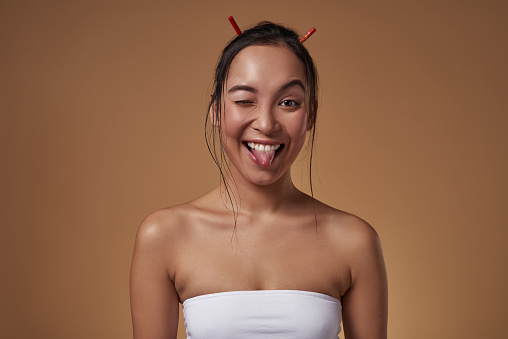 Joking fashionable asian girl winking and sticking out tongue. Pretty young brunette slim woman wearing tank top. Female beauty. Isolated on orange background. Studio shoot. Copy space