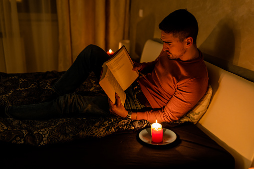 A young man is lying in bed and reading a book during an electricity crisis causing a blackout under the lit candles.