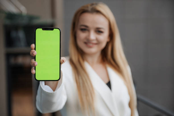 istock Blond smiling woman at oversized white jacket showing phone screen with chromakey on city background 1454367200