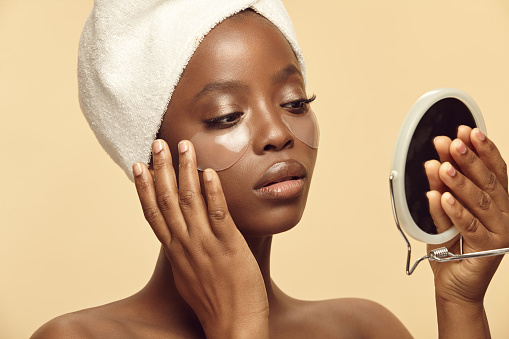 African ethnicity woman in white head towel applying eye patches on beige background while looking at the mirror. Under eye masks for puffiness, wrinkles, dark circles. Eye patches concept.
