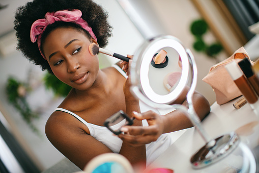 Young woman applying blusher on her cheeks while sitting in front of the mirror in her home.