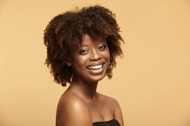 Beautiful portrait of cheerful smiling African American ethnicity woman with a clean healthy skin and afro style hairstyle on a beige isolated background. Beautiful portrait of cheerful smiling African American ethnicity woman with a clean healthy skin and afro style hairstyle standing on a beige isolated background. strapless stock pictures, royalty-free photos & images