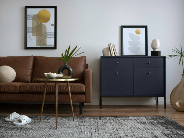 istock Interior design of harmonized living room with mock up poster frame, brown sofa, navy commode, coffee table, carpet, leaf in vase, and personal accessories. Creative home decor. Template. 1454373205