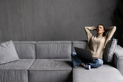 Business young woman sitting on sofa with laptop raising her arms above her head stretching after sitting for a long time at work.