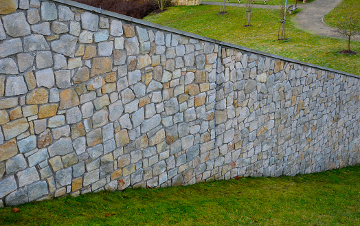 The plank fence follows the sandstone-lined wall that is cracking in several places, the movements of the terrain and the vibrations of the highway car traffic. grassy slope and high wall sloping down, thermic