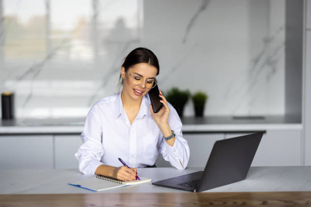 istock Smiling business woman working with laptop while talking with smartphone in the kitchen at home. 1454374471