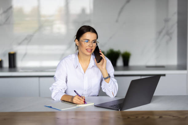 istock Smiling business woman working with laptop while talking with smartphone in the kitchen at home. 1454374492
