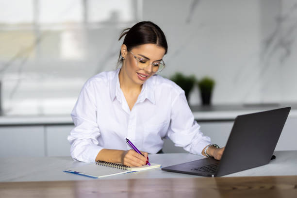 istock Young woman sit at table with laptop in kitchen holds pen, take notes on paper, writes letter. 1454374891