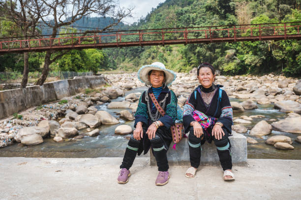 front view on two indigenous woman from black hmong tribe sitting on bridge and looking to camera in the sa pa mountain region in north vietnam