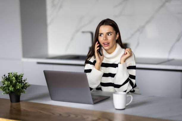 istock Young woman sits at the kitchen table using a laptop and talking on phone 1454375500