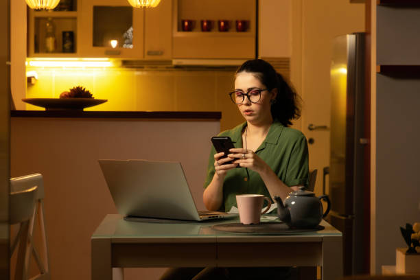istock Thoughtful young Caucasian woman wearing glasses uses smartphone while sitting at table with laptop. Concept of freelancing and social media. 1454375644