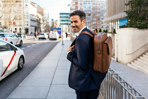 Back view of mature male entrepreneur with leather backpack walking along street and commuting to work in morning