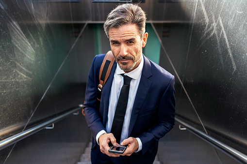 High angle of confident mature male entrepreneur in suit and with mobile phone standing on staircase of underpass and looking at camera