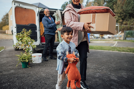 A shot of a mature couple wearing casual clothing unloading boxes and furniture from the rear of a moving van, ready to move in to their new bungalow together. They are with their young grandson.