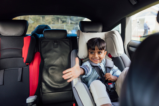 Taken from inside of a car, a front-view shot of a young boy sitting in his car seat with the safety harness on. He is looking away from the camera and is ready to go on a car journey.