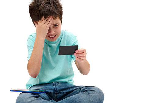 Dissatisfied Caucasian teenage boy in blue t-shirt, looking at his credit card, expressing disappointment and frustration while failing transaction, isolated on white background. Unsuccessful payment