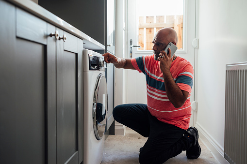 A close-up shot of a mature man crouching down near his washing machine, he is calling an appliance repair service on his smartphone at his home in the North East of England.