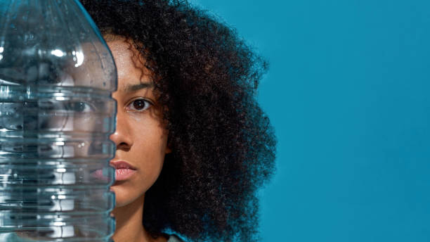 Partial image of young black girl holding plastic bottle and looking at camera. Ecology safety and protection. Waste disposal and recycling. Isolated on blue background. Studio shoot. Copy space