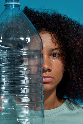 Cropped image of young black girl holding plastic bottle and looking at camera. Ecology safety and protection. Waste disposal and recycling. Environmental sustainability. Blue background. Studio shoot
