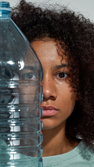 Partial image of black girl holding plastic bottle and looking at camera. Ecology safety and protection. Waste disposal and recycling. Environmental sustainability. White background. Studio shoot